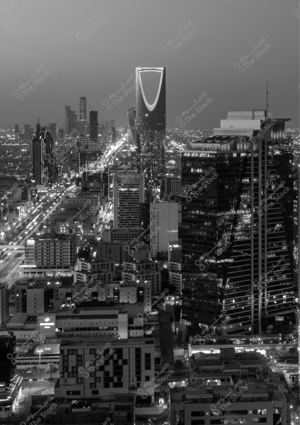 The image shows a nighttime view of Riyadh, Saudi Arabia. In the center, the iconic Kingdom Tower stands out with its distinctive illuminated design. It is surrounded by modern buildings and the lights emanating from the streets and intersections, giving the city an advanced urban feel. The skyline reveals city details under a cloudy sky.