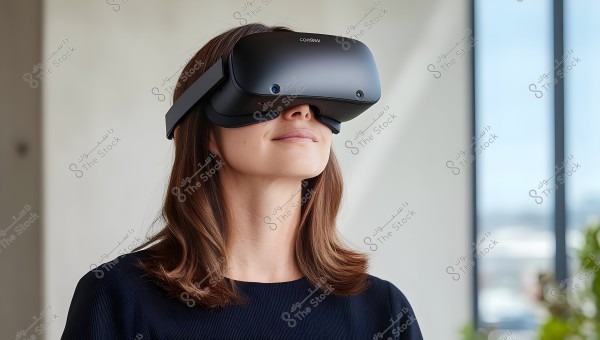 A woman wearing black virtual reality goggles, standing in a sunlit indoor space, wearing a black blouse. Her hair is long, brown and straight. In the background, there is a large glass window with some green plants in the corner.