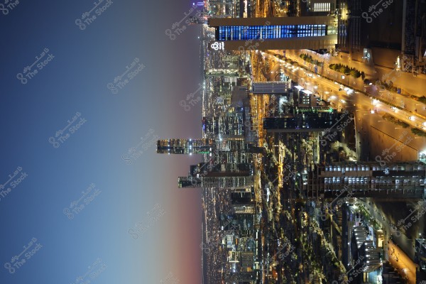 لقطة ليلية لمدينة مضاءة بالأضواء المتلألئة. المباني الشاهقة تصطف على طول الطريق المضاء, مع حركة مرور متفرقة. السماء تتدرج من ألوان الغروب الكحلي إلى الوردي, مما يخلق مشهدًا ساحرًا.