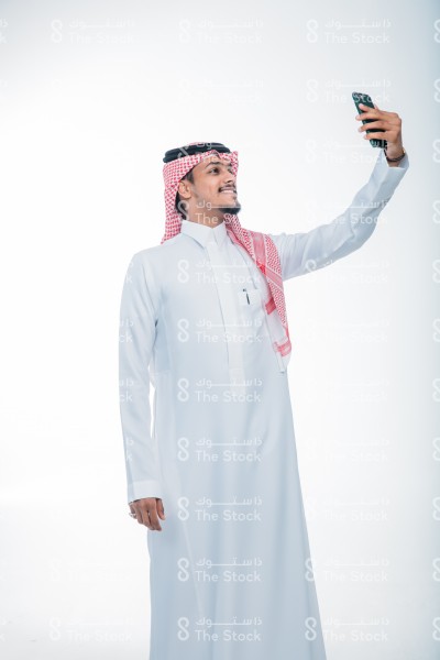 بورتريه رجل سعودي أمام خلفية بيضاء يرتدي الثوب ويبتسم. صورة شخصية لرجل سعودي مبتسم، نظر إلى الجوال ،يقوم بالتقاط صور سيلفي ، يستخدم وسائل التواصل الاجتماعي ، توثيق لحظة سعيدة ، صور تذكارية