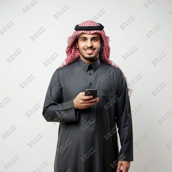Image shows a man wearing a dark thobe and a red and white ghutra. He stands against a white background holding a smartphone in his hands and smiling.