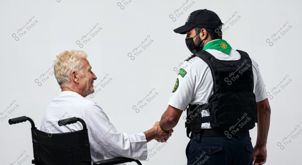 An image of an elderly man sitting in a wheelchair wearing a white shirt, shaking hands with a security officer wearing a uniform consisting of a white shirt, protective vest, and black cap. The two men are engaging in a friendly interaction against a white background. The security officer\'s uniform displays Arabic text, suggesting the setting may be in Saudi Arabia.