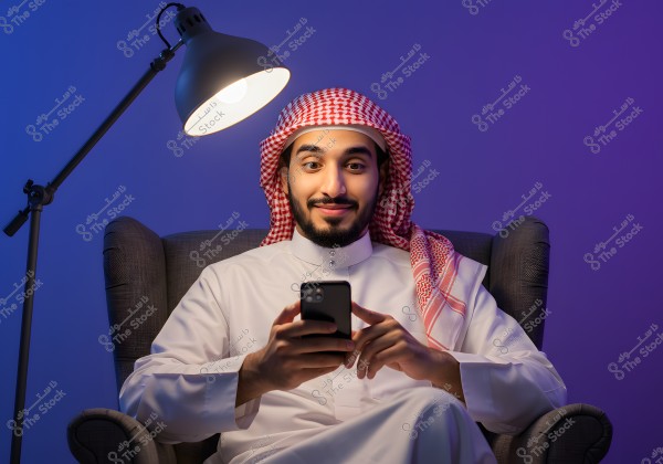 A portrait of a man sitting on a comfortable chair under the light of a modern lamp. The man is wearing a white thobe and a red and white ghutra, indicating traditional Saudi attire. He is smiling and looking at a smartphone in his hands. The background is blue, creating a calm atmosphere.