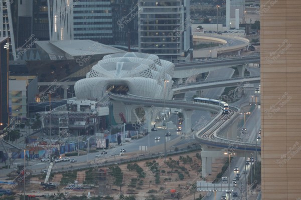 منظر للمدينة يظهر فيه مبنى محطة مترو حديث ذو تصميم معماري فريد باللون الأبيض, مع خطوط أنيقة وإنحناءات. يحيط بالمبنى جسور وطرق سريعة مزدحمة بالسيارات, مع وجود قطار يتجه نحو المحطة. تتواجد في الخلفية مباني شاهقة ومعمار حديث.