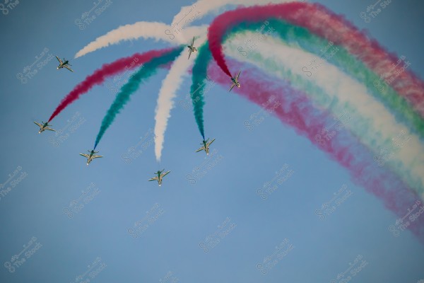 Aircraft in an aerial formation release colored smoke trails in red, green, and white against the blue sky. The planes are arranged in a geometric pattern, leaving vibrant trails behind.