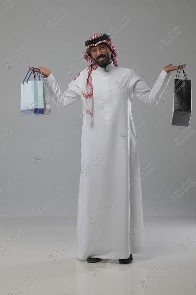A portrait of a man standing in a studio with a gray background, wearing a traditional white Saudi thobe and a red and white shemagh, with glasses on. The man appears to be smiling and holding shopping bags, one in each hand.