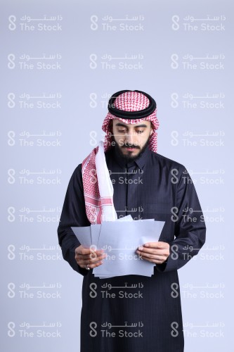 بورتريه رجل سعودي يرتدي الزي السعودي التقليدي يحمل بكلتا يديه مجموعة من الأوراق وينظر إليها، معلم سعودي ينظر لأوراق الأختبار