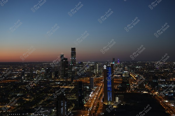 A panoramic view of Riyadh city at dusk, displaying the illuminated cityscape with buildings and lit streets. The high-rise towers and modern architecture are prominent, with beautiful gradient colors in the sky ranging from light orange to dark blue.