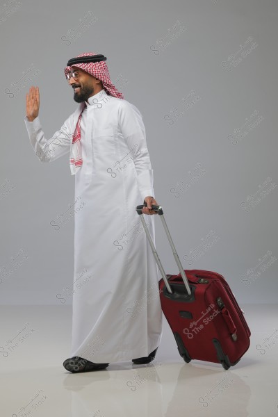 Image of a man wearing a white thobe and a red and white checked headscarf (ghutra) with an agal, waving goodbye while pulling a red wheeled suitcase. He is smiling and standing against a gray background.