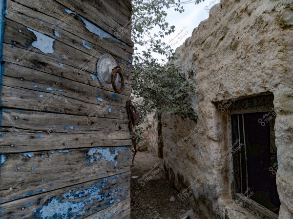 An image of a narrow alley between old stone walls. The left wall features a weathered wooden door with faded blue paint and a rusty metal handle. The right wall shows an open window and a small entrance. A climbing plant extends across the stone wall.