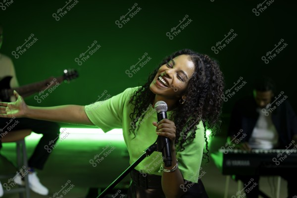 امرأة تغني بحماس وهي تمسك الميكروفون, يشع خلفها ضوء أخضر, ويمكن رؤية موسيقين يعزفون على الجيتار والبيانو في الخلفية.