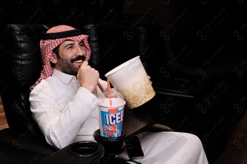 شاب سعودي يرتدي الزي السعودي التقليدي يستمتع بمشاهدة الفيلم في صالة السينما ويحمل علبة الفشار بينما يظهر أمامه أحد المشروبات على الطاولة