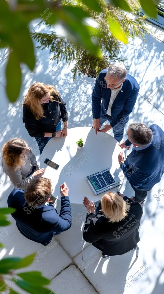 A group of people gathered around a round table in a sunlit outdoor space. Everyone is wearing formal business attire with a laptop and a smartphone on the table. Green trees surround the area, adding a touch of nature to the environment.
