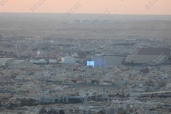 The image shows an aerial view of the city with dense buildings and streets of Riyadh. The cityscape dominates the image with an open space in the background that includes white buildings, a dark spherical structure, and a large illuminated building at the front. In the middle, there is a lit Ferris wheel in what appears to be an amusement area. The sky is tinged with shades of orange, indicating either sunrise or sunset.