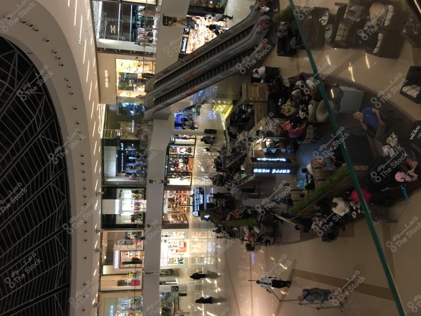 An image of an indoor shopping mall featuring various stores and a café in the center. People are seen shopping and walking around while others are seated at the café. The area is well-lit with a modern design, including escalators connecting the floors.