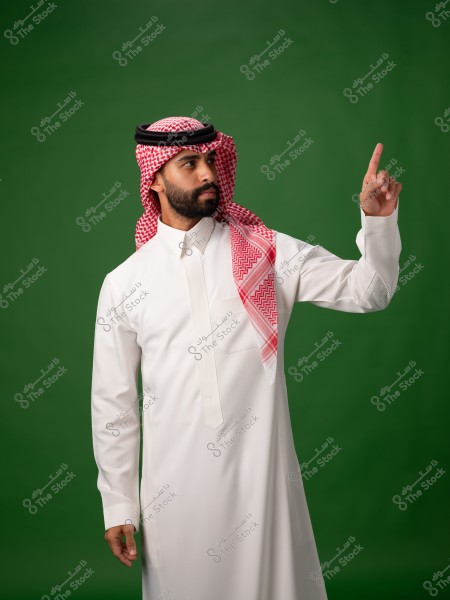 Image of a man wearing traditional white attire and a red checkered headscarf (ghutra) with a black band (agal) standing against a green background. He appears to be pointing upwards with his finger.