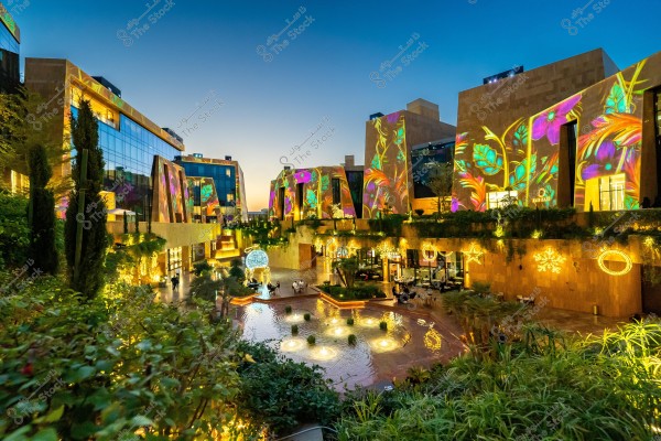 An image of a modern shopping complex lit up with colorful lights in the evening. The facade is decorated with illuminated floral designs in vibrant colors, including blue and pink. There\'s also a small water pond with lit fountains, and trees and plants in the foreground and background are adorned with twinkling decorative lights. The building facades feature large glass windows reflecting the surrounding lights.