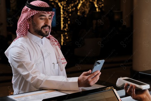 شاب سعودي في استقبال الفندق ، ويقوم بتعابير مختلفة باستخدام الجوال  ، ويقوم بدفع عبر جهاز الدفع الالكتروني