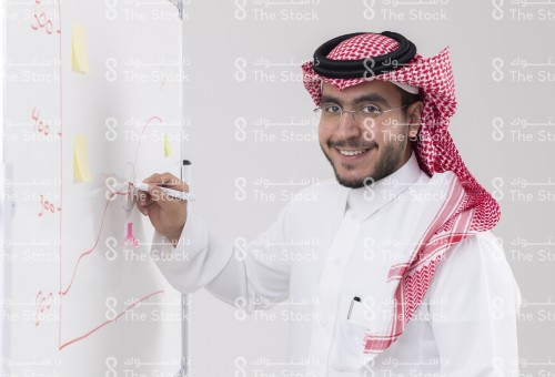 شاب سعودي مبتسم يرتدي الزي السعودي التقليدي يقوم بالرسم البياني على السبورة، رجل يكتب على اللوح