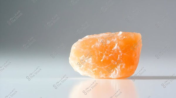 Image showing a piece of natural pink salt placed on a smooth, light gray surface. The piece is irregularly shaped and features clear details with a slight reflection on the surface. The background is plain and simple, highlighting the beauty and details of the salt.