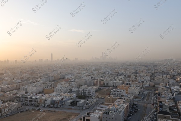 The image shows an aerial view of a city with sprawling multi-story residential buildings under a clear sky. In the background, a cluster of skyscrapers and business centers loom on the horizon, indicating a modern Middle Eastern city. The sky appears bright with soft sunlight almost covering the horizon.
