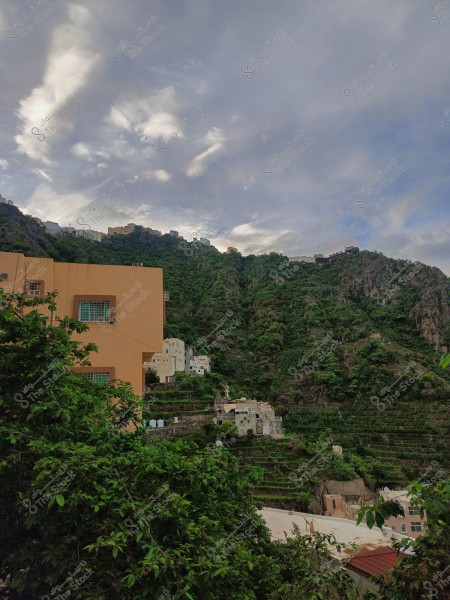 منظر طبيعي يظهر سلسلة من التلال الخضراء, تنتشر عليها المنازل البيضاء والبنية المتناغمة مع البيئة, مع سماء ملبدة بالغيوم في الخلفية. في المقدمة, يظهر مبنى بلون برتقالي وأشجار خضراء.