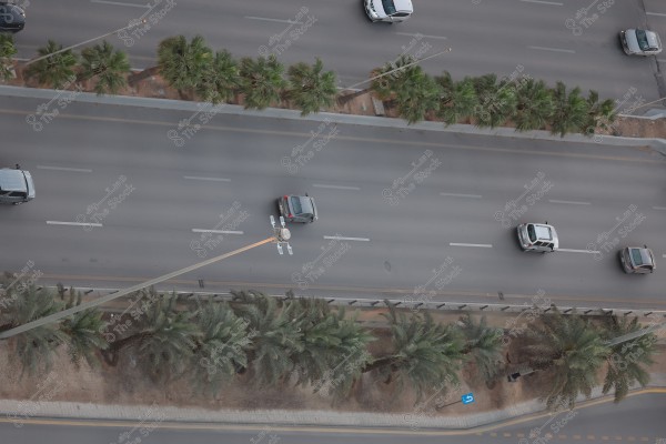 An aerial view of a highway with six lanes, divided into three lanes in each direction. Several cars are seen driving in both directions. A row of green palm trees separates the two directions, with more palm trees lining the side of the road. Streetlights are distributed along the road.
