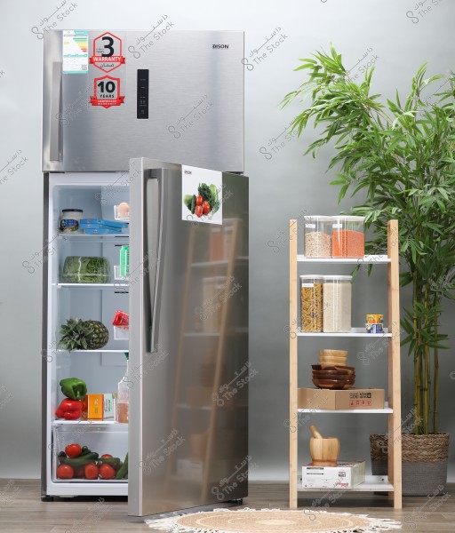 Image of a modern kitchen featuring a silver refrigerator with an open door showing several shelves filled with vegetables like tomatoes, green peppers, and pineapple. Next to the refrigerator is a wooden shelf with transparent storage containers filled with dry ingredients like pasta and rice, along with wooden bowls. There is a round rug on the floor and a large plant in the background corner.