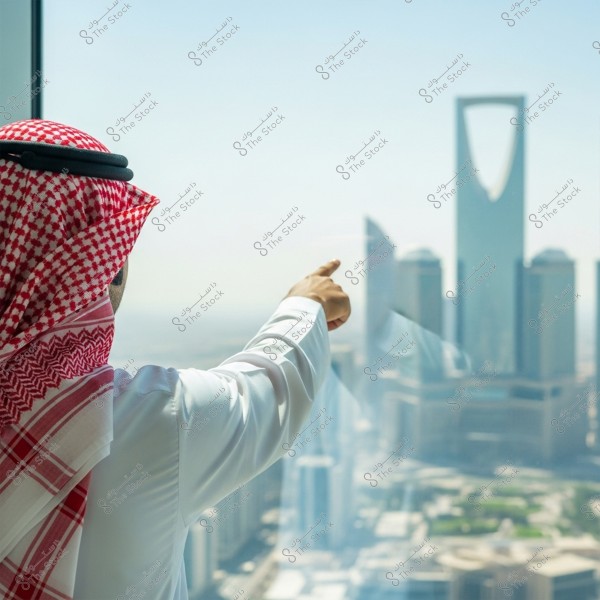 ** رجل يرتدي الزي التقليدي السعودي, يشير بيده نحو برج مميز في الأفق. الزي يتكون من شماغ أحمر وأبيض وعقال, بالإضافة إلى ثوب أبيض. في الخلفية, يظهر أفق مدينة الرياض بوضوح مع برج المملكة البارز.\r\n\r\n**