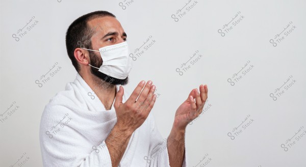 Image of a man wearing a white Ihram, standing with his hands raised as if in prayer or supplication. He is also wearing a face mask. The background is entirely white, emphasizing the subject\'s details.