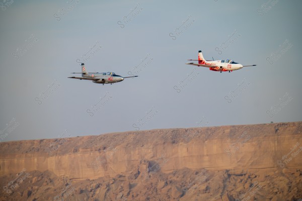 Two military aircraft flying in the clear sky above a desert landscape. The aircraft on the left is gray with the number \"1402,\" while the aircraft on the right is white with a red design and the number \"173.\" In the background, there is a large rocky cliff resembling desert mountains.