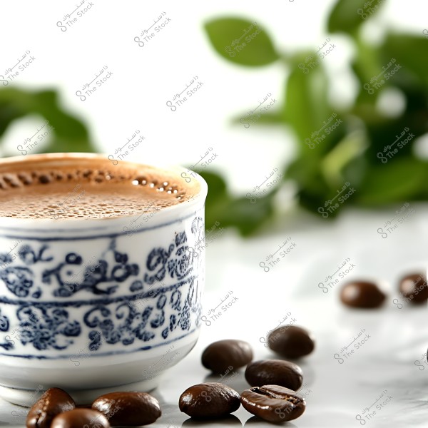 A blue and white decorated coffee cup filled with fresh coffee, featuring bubbles on the surface, placed on a white surface. Scattered around the cup are coffee beans. In the background, blurry green plant leaves can be seen, adding a natural feel.