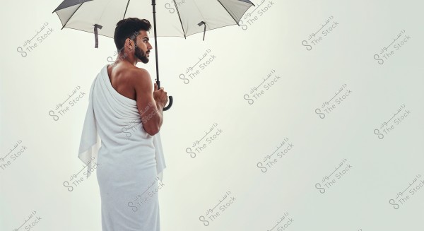 A photo of a man wearing a white draped garment, seen from behind, holding an open umbrella. The background is light and plain, highlighting the details of the garment and umbrella. The white draped attire resembles traditional Arab or Roman styles.