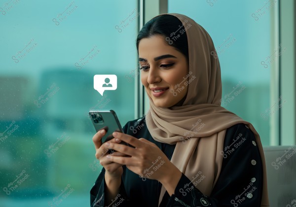 A woman wearing a beige hijab and a black abaya is sitting near a window. She is holding a mobile phone and smiling as she looks at it, with a chat icon displayed beside her.