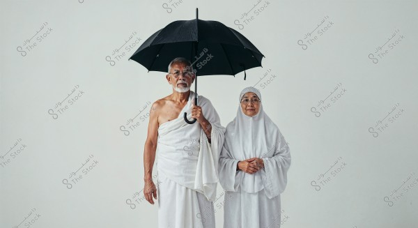 The image shows a man and a woman standing together. The man is wearing the traditional white Ihram clothing, and the woman is dressed in a white hijab and gown. The man is holding a black umbrella. The white background highlights their white attire.