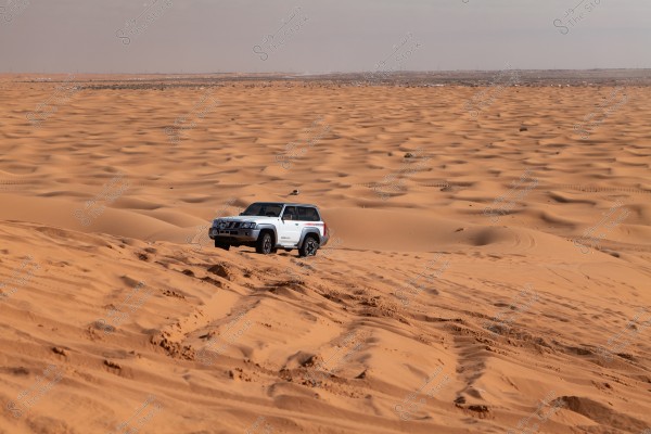 A white SUV driving through the sand dunes in an open desert. The sky is clear and the sands stretch to the horizon, suggesting isolation and the beauty of the desert landscape.