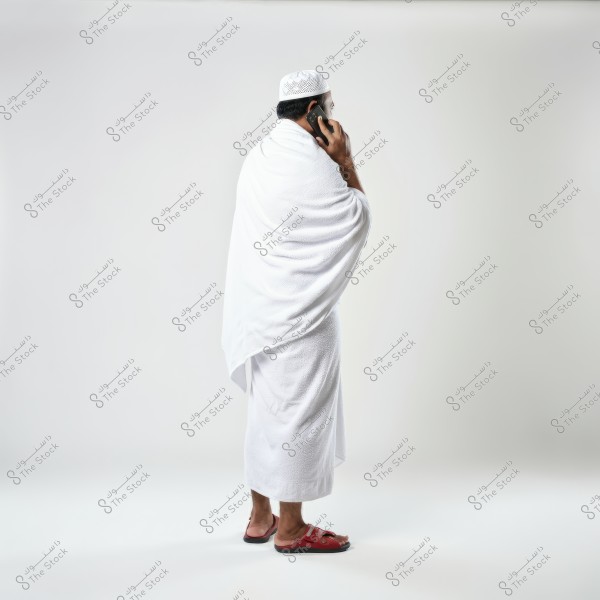 The image depicts a man wearing the traditional white Ihram clothing consisting of two pieces, with a white cap on his head. He is standing in a side view and talking on a mobile phone, wearing red sandals. The background is white and clean.