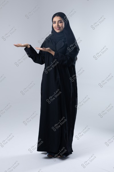 A woman wearing a black abaya and hijab stands with a slight smile on a simple gray background. She holds her hands up in a welcoming gesture. The image reflects the cultural aspect of traditional attire.