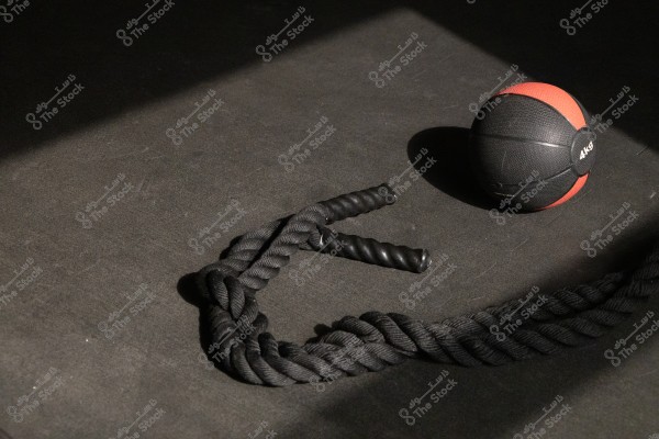 A black and red medicine ball weighing 4 kg is placed on a dark floor next to a long black exercise rope with wrapped handles. Lighting highlights parts of the ball and rope, emphasizing the textures and shadows.