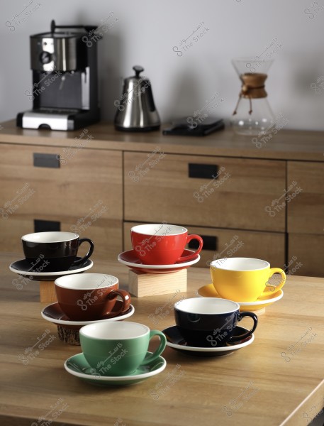 Six colorful coffee cups on a wooden table with a coffee machine and brewing tools in the background.