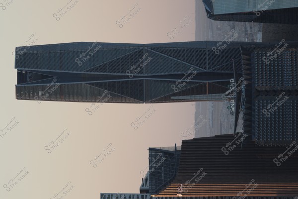 منظر لناطحة سحاب حديثة بتصميم هندسي معقد في منطقة حضرية. يحتوي المبنى على هيكل زجاجي ومعدني ويظهر وسط مباني أخرى متنوعة في التصميم والارتفاع. توجد علامة مكتوبة بالأحرف الإنجليزية على المبنى "SNB". السماء في الخلفية تبدو صافية.