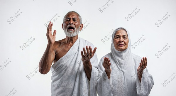 Image of an elderly man and woman in a prayer position, wearing ihram clothing. The man on the left wears a white izar, while the woman beside him on the right wears a white hijab and an embroidered robe. The background is white.