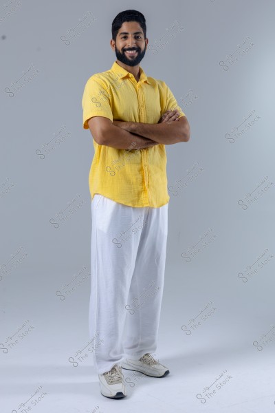 A portrait of a man standing and smiling, wearing a yellow shirt and white loose-fitting pants with white sneakers. The man is standing with his arms crossed over his chest, appearing in a relaxed setting.