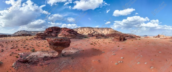 A wide desert landscape featuring red sand and large, uniquely shaped rock formations. White clouds float in the clear blue sky, casting light shadows on the ground. The terrain varies between prominent rocks and flat areas.