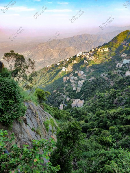 A breathtaking natural landscape showing a series of mountains covered in dense green vegetation with scattered houses on their slopes. The sky is clear and blue above, with the mountains extending to the horizon appearing in a light purple hue due to distance.