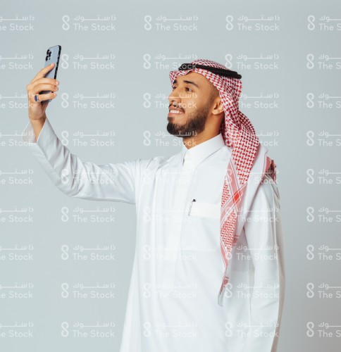 بورتريه رجل سعودي مبتسم يرتدي الزي السعودي التقليدي يرفع الجوال لالتقاط السيلفي، شاب يأخذ صورة شخصية بالهاتف المحمول