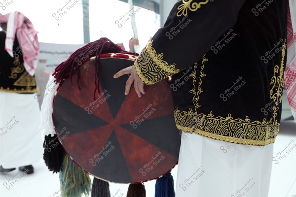 A person wearing traditional clothing adorned with gold embroidery holds a large drum. Another side of a person wearing a shemagh and ghutra is visible. The drum is decorated with intersecting lines and multiple colors. The image appears to depict a cultural or heritage event.