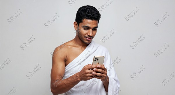 A portrait of a man wearing a white garment similar to ihram clothing, looking at a gold smartphone in his hands. The background is light gray, and the man appears focused while using the phone.