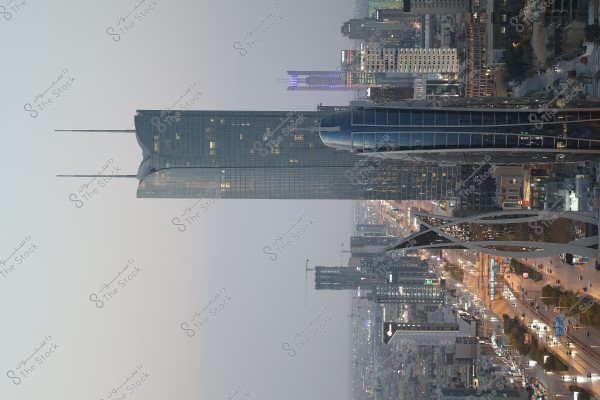 View of prominent skyscrapers in a modern city during dusk. The buildings feature contemporary architectural designs and illuminated lights. In the foreground, a busy street with cars and street lighting creates an advanced urban atmosphere.