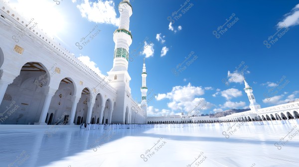 Image depicting the Al-Masjid an-Nabawi in Medina, Saudi Arabia. The mosque\'s Islamic architectural design is highlighted with decorative white arches and prominent tall minarets. The sky is blue and clear with a few clouds, providing a serene and majestic view of the sacred place. Worshippers can be seen in the courtyard.